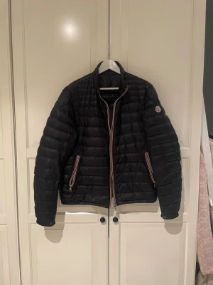 Moncler daniel dunjacka - Snygg mörkblå dunjacka från Moncler. Storlek 5 som motsvarar L/XL. Jackan har två framfickor med dragkedja och Moncler-logga på ärmen. Dragkedjorna har rödvita detaljer som ger en sportig touch. Perfekt för kalla vinterdagar eller kyligare vårdagar.