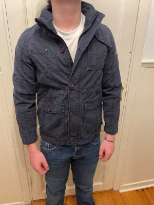 Mörkblå casual jacka med knappar - Snygg mörkblå jacka med hög krage och flera fickor framtill. Jackan har knappstängning och är tillverkad i ett slitstarkt bomullsmaterial. Passformen är normal och jackan har långa ärmar, perfekt för vardagslooken.