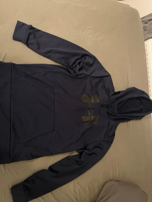 Mörkblå hoodie från Under Armour - Snygg mörkblå hoodie från Under Armour med stor logga på bröstet. Hoodien har en klassisk känguruficka framtill, justerbar huva med dragsko och mjukt fleecefoder på insidan. Perfekt för dig som gillar sportig och avslappnad stil.