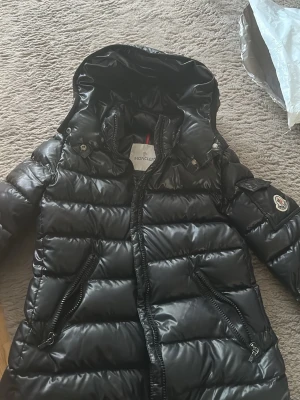 Svart dunjacka från Moncler med huva - Snygg svart dunjacka från Moncler med glansig finish, avtagbar huva och två dragkedjefickor framtill. Klassisk Moncler-logga på ärmen och quiltad design. Perfekt för kalla vinterdagar. Xxs 152