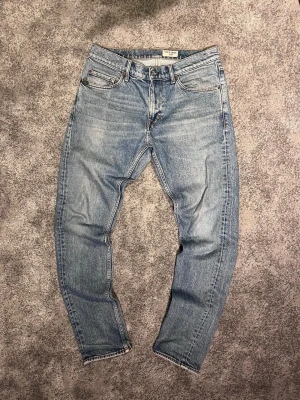 Ljusblå raka jeans från Nudie Jeans - Säljer ett par ljusblå raka jeans från Nudie Jeans med klassisk femficksmodell och knappgylf. Jeansen har en avslappnad passform och är tillverkade i 100% bomull. Perfekta för en casual och trendig look.