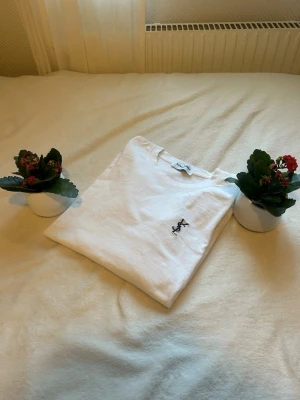 Vit Saint Laurent t-shirt - Säljer en clean vit t-shirt från Saint Laurent med klassisk crewneck och diskret svart YSL-logga broderad på bröstet. T-shirten är i mjuk bomull och har en enkel, stilren vibe som passar till allt.