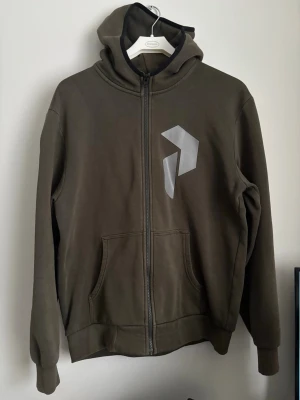 Olivgrön hoodie med dragkedja Peak Performance  - Olivgrön hoodie med dragkedja framtill och stor ficka på magen. Har en stor huva och ett stilrent, geometriskt tryck i grått på bröstet. Tillverkad i mjukt sweatshirtmaterial, perfekt för en avslappnad look.