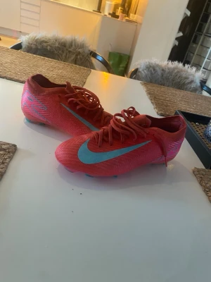 Nike Mercurial röda fotbollsskor - Säljer ett par Nike Mercurial PRO version fotbollsskor i rött med ljusblå swoosh och detaljer. Skorna har en smal och aerodynamisk form, tillverkade i syntetmaterial för bra grepp och känsla på planen. Perfekta för dig som vill ha snabba fötter och snygg stil på matchen.