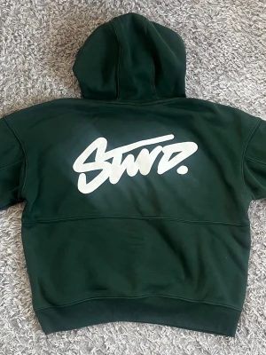 Mörkgrön Stvrd hoodie med tryck - Mörkgrön hoodie från Stvrd med stor vit logga på ryggen och mindre logga på bröstet. Klassisk känguruficka framtill och justerbar huva med dragsko. Tillverkad i mjukt bomullsmaterial som är perfekt för chill dagar.