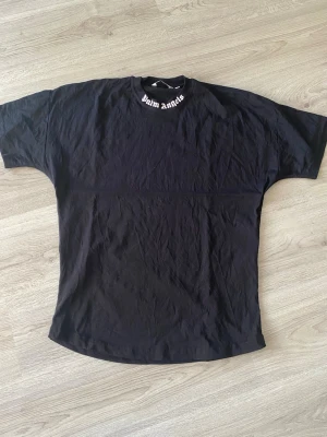Svart Palm Angels t-shirt med tryck - Cool svart t-shirt från Palm Angels med stort vitt logotryck på ryggen och mindre text på kragen. Oversized passform och tillverkad i mjuk bomull. Perfekt för dig som gillar streetwear och vill sticka ut med en ikonisk design.