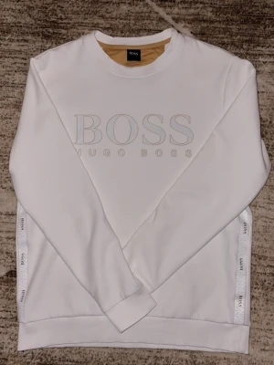 Hugo Boss tröja - Snygg vit sweatshirt från Hugo Boss med stort BOSS-tryck på bröstet och diskreta logoband längs sidorna. Tröjan har rund halsringning och ribbade muddar vid ärmar och nederkant. Perfekt för en clean och stilren look.