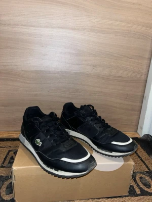 Svarta Lacoste sneakers med vit sula - Storlek 43.Snygga svarta sneakers från Lacoste med vit sula och grönt krokodilemblem på sidan. Skorna har en klassisk låg profil, svarta skosnören och detaljer i både mocka och textil. Perfekta för en clean och sportig look.