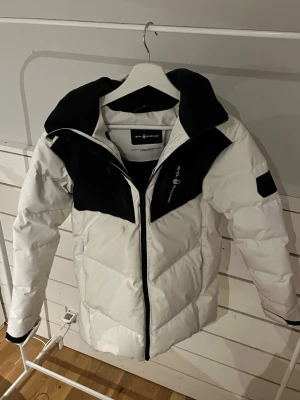 Vit och svart pufferjacka Sail Racing - Säljer en riktigt snygg vit och svart pufferjacka från Sail Racing. Jackan har huva, dragkedja framtill och coola logodetaljer på bröstet och huvan. Fodrad insida och flera fickor med dragkedja. Perfekt för kalla dagar när du vill hålla stilen.