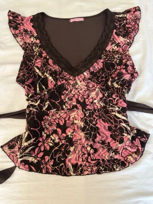 Blommig volangblus med spetskant - Säljer en blommig blus i rosa och svart med volangdetaljer på axlarna och djup v-ringning med svart spetskant. Blusen har omlottkänsla och knytband i midjan för en feminin siluett. Perfekt för dig som gillar romantiska och unika toppar.