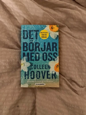 Det börjar med oss av Colleen Hoover - En roman med blått omslag och stora blommor i vitt och orange. Titeln 'Det börjar med oss' står i stora bokstäver och författaren är Colleen Hoover. Boken är tryckt på papper och ges ut av Lovereads.