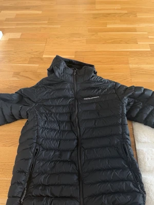 Svart dunjacka från Peak Performance - Snygg svart dunjacka från Peak Performance med huva och dragkedja framtill. Jackan har två sidofickor med dragkedja och diskret logga på bröstet. Lättviktigt material och quiltad design som håller värmen under kalla dagar.
