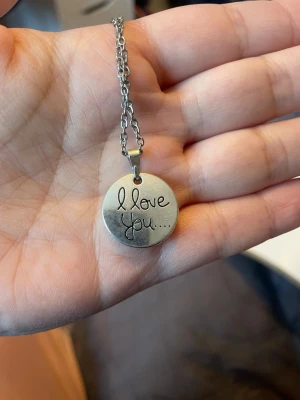 Silverfärgat halsband med textberlock - Snyggt halsband i silverfärgad metall med en rund berlock där det står 'I love you...' ingraverat. Kedjan är enkel och stilren, och berlocken har en matt finish. Perfekt accessoar för dig som vill bära ett personligt budskap nära hjärtat.