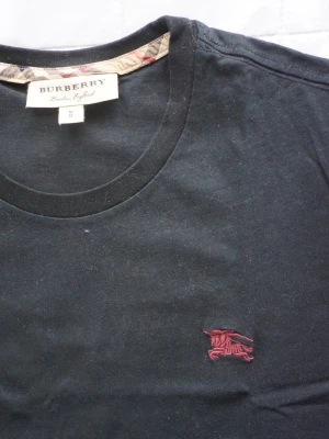 Burberry | Svary Tshirt - Klassisk svart t-shirt från Burberry med rund halsringning och diskret röd broderad logga på bröstet. T-shirten är tillverkad i mjuk bomull och har korta ärmar. Perfekt för en stilren och enkel look. Helt oanvänd, passar åt slim hållet! 