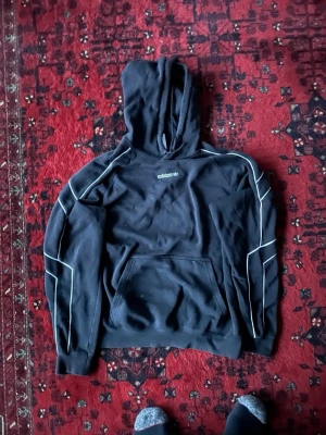 Svart hoodie från Adidas med vita detaljer - Säljer en svart hoodie från Adidas med vita kontrastsömmar längs ärmar och axlar. 