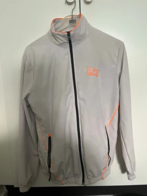 Vit dress från EA7 Emporio Armani - Snygg dress  från EA7 Emporio Armani med orange detaljer och logga på bröstet. Jackan har hel dragkedja framtill, två fickor med svarta dragkedjor och elastiska muddar. Perfekt för dig som vill ha en stilren och sportig look. (Byxor och tröja)