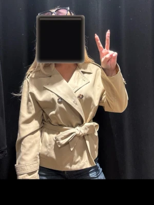 Kort beige trenchcoat stradivarius - Stilren beige trenchcoat i kortare modell med dubbelknäppning, bred krage och knytskärp i midjan. Jackan har axelklaffar och dekorativa knappar vid ärmslut. Använd fåtal gånger och har för många jackor.
