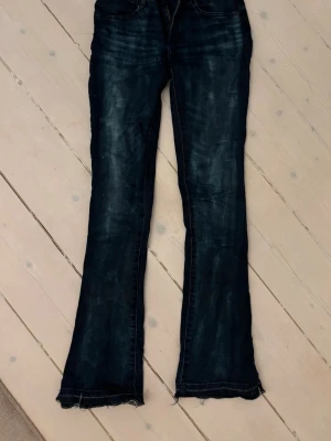 Mörkblå bootcut jeans med rå kant - Säljer ett par mörkblå bootcut jeans med slitningar och råa, fransiga benslut. Jeansen har klassisk femficksdesign och markerade sömmar. Perfekta för en avslappnad och trendig look.