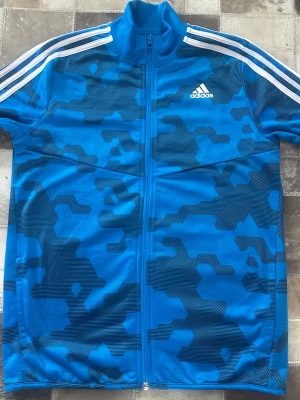 Blå träningsjacka/kofta  från Adidas - Snygg blå träningsjacka från Adidas med vit logga på bröstet och klassiska vita ränder på axlarna. En  hel dragkedja framtill. Tillverkad i ett lätt och funktionellt material, perfekt för träning eller sport. Helt ny oanvänd