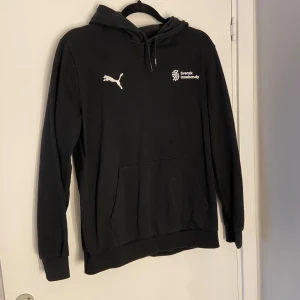 Svart hoodie från Puma Svensk Innebandy - Svart hoodie från Puma med tryck av Svensk Innebandy på bröstet. Klassisk känguruficka framtill, justerbar huva med snören och ribbade muddar. Tillverkad i mjukt bomullsmaterial för en skön och avslappnad stil.