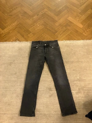 Svarta slim fit jeans från Nudie - Säljer ett par svarta slim fit jeans från Nudie Jeans med klassisk femficksmodell och diskreta slitningar. Jeansen har normal midja, dragkedjegylf och är tillverkade i stretchigt denim för skön passform. Perfekta till en avslappnad streetstyle-look. Grim Tim 30-30