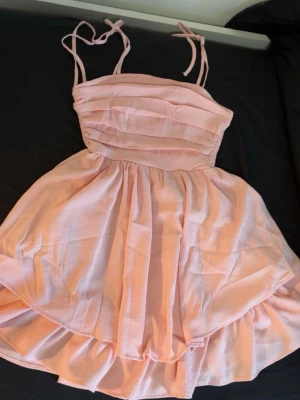 Ljusrosa kort klänning  - Söt ljusrosa kort klänning med tunna axelband som knyts upptill. Klänningen har ett rynkat liv och volang i kjolen för en romantisk look. Tillverkad i ett lätt och luftigt tyg, perfekt för varma dagar.