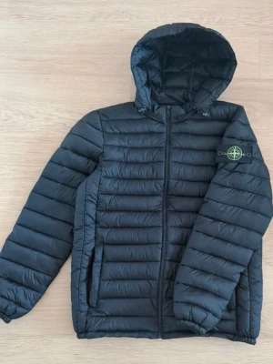 Svart pufferjacka från Stone Island - Säljer en svart pufferjacka från Stone Island med huva och dragkedja framtill. Jackan har två sidofickor med dragkedja och den klassiska Stone Island-loggan på vänster ärm. Quiltad design och elastiska muddar vid ärmsluten. Perfekt för kalla vinterdagar. Strl M helt ny