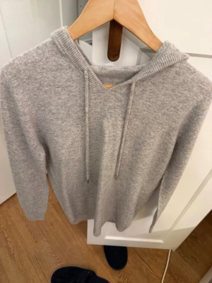 Cashmire hoodie - Mysig ljusgrå hoodie med huva och dragsko. Tillverkad i mjukt stickat material som känns skönt mot huden. Hoodien har en enkel och stilren design utan tryck eller logga, perfekt för en avslappnad look. Inget brand