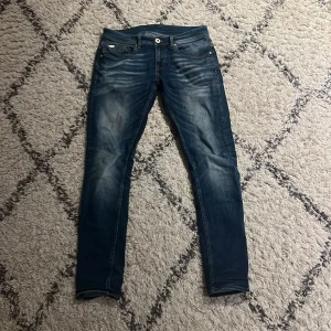 Blå slim fit jeans med slitningar - Säljer ett par blå slim fit jeans med snygga slitningar och tvättade detaljer. Klassisk femficksmodell med knappgylf och läderpatch bak i midjan. Jeansen har smal passform och är perfekta för en avslappnad men trendig look. 29W 32L