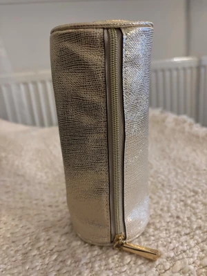 Cylindrisk guldig necessär med dragkedja - Säljer en unik cylindrisk necessär i skimrande guldigt material med struktur. Necessären har en dragkedja i matchande guld och en stilren look. Perfekt för dig som vill sticka ut med accessoarer i metallic och gillar modern design.