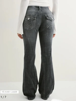 Grå bootcut jeans från True Religion - Säljer ett par grå bootcut jeans från True Religion med tydliga vita kontrastsömmar och stora fickor bak med lock och knappar. Jeansen har en klassisk låg midja och är tillverkade i denim med en lätt tvättad look. Perfekta för en avslappnad men trendig stil.
