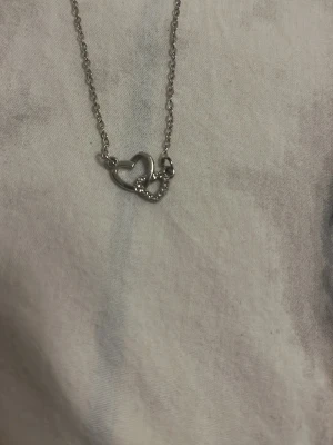 Silverhalsband med dubbla hjärtan - Snyggt halsband i silverfärg med två sammanlänkade hjärtan, där det ena hjärtat är dekorerat med små gnistrande stenar. Kedjan är tunn och elegant, perfekt för att ge en subtil touch till din outfit. Passar dig som gillar stilrena och romantiska smycken. I mycket bra skick används 2 gånger på bröllop 