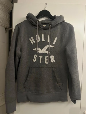 Grå hoodie från Hollister - Säljer en grå hoodie från Hollister med stor magficka och vit broderad logga och text på bröstet. Hoodien har dragsko i huvan och ribbade muddar vid ärmar och nederkant. Perfekt för en avslappnad och stilren look.