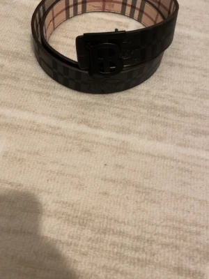 Svart rutigt skärp från Burberry - Snyggt svart skärp från Burberry med rutigt mönster och en stor B-logga på spännet. Skärpet är gjort i skinn och har gulddetaljer. Perfekt accessoar för att lyfta din outfit.