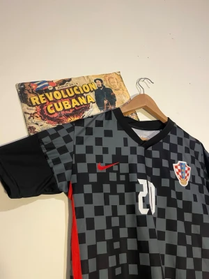 Brate moi! Kroatien fotbollströja Petković #20 - Snygg svart och grå rutmönstrad fotbollströja från Nike med Kroatiens landslagsemblem på bröstet och Petković samt nummer 20 på ryggen. Tröjan har röda detaljer och är tillverkad i ett lätt, ventilerande material. Perfekt för match eller träning.