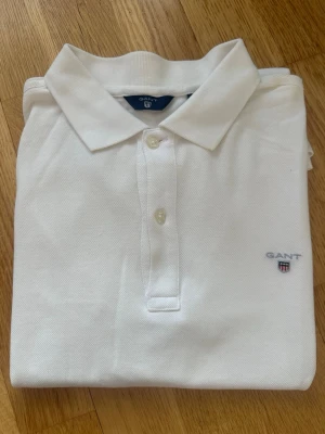 Vit pikétröja från GANT - Klassisk vit pikétröja från GANT med krage och knappar framtill. Tröjan har diskret GANT-logga broderad på bröstet och är tillverkad i ett mjukt bomullsmaterial. Perfekt till en clean och stilren look.