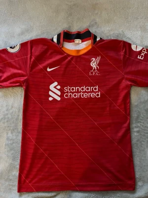 Liverpool matchtröja Nike Salah 11 - Säljer en röd Liverpool FC matchtröja från Nike med tryck av M. Salah och nummer 11 på ryggen. Tröjan har klubbmärke, sponsorlogga och Premier League-märken på ärmarna. Klassisk krage med svarta och orange detaljer. Perfekt för fotbollsfans!