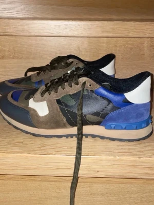 Valentino camo sneakers i mocka och läder - Snygga sneakers från Valentino med camomönster i grönt, blått och svart. Skorna har detaljer i mocka och läder, blå hälkappa med nitar och olivgröna skosnören. Yttersulan är beige och svart, och insidan är svart. Perfekta för dig som gillar streetstyle och unika detaljer.