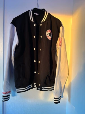 Converse svart och vit bomberjacka - Svart och vit bomberjacka från Converse med klassisk college-look. Jackan har vita ärmar, svart kropp, broderad Converse-logga på bröstet och tryck på ryggen. Ribbstickade muddar och krage med ränder. Perfekt för en sportig streetstyle. Fläckar finns, se bilder. Storlek xxl men är som en M