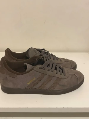 Bruna Adidas Gazelle sneakers - Säljer ett par bruna Adidas Gazelle sneakers i mocka med klassiska tre ränder på sidan och guldfärgad 'GAZELLE'-text. Skorna har lågt skaft, matchande bruna skosnören och en mörkbrun sula. Perfekta för dig som gillar retrostil och streetwear.