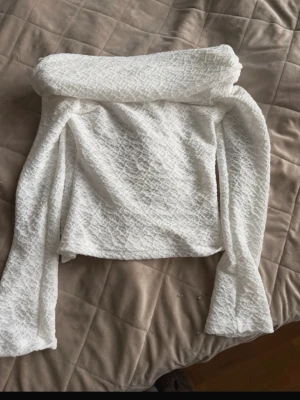 Vit offshoulder topp i strukturtyg - Trendig vit offshoulder topp med lång ärm i ett strukturerat tyg som ger en unik känsla. Toppen är croppad och har en snygg, figurnära passform. Perfekt för dig som vill sticka ut med en stilren men ändå cool look.