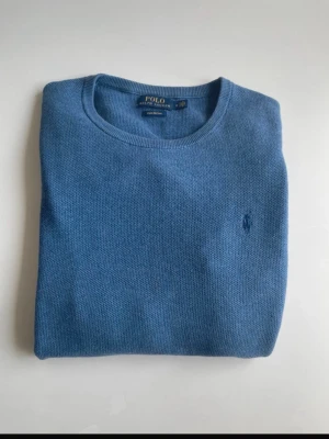 Blå stickad tröja från Polo Ralph Lauren - Snygg blå stickad tröja från Polo Ralph Lauren med rund halsringning och diskret broderad logga på bröstet. Tröjan har långa ärmar och ribbade muddar vid ärmslut och nederkant. Perfekt för lager på lager och stilrena outfits.