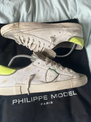 Vita Philippe Model sneakers med neonhäl - Säljer ett par vita Philippe Model sneakers i läder med coola neon-gula detaljer på hälen och grönt kantband vid loggan. Klassisk låg modell med vita snören och unik sula där det står PHILIPPE MODEL. Perfekta för dig som gillar streetstyle och exklusiva märken.