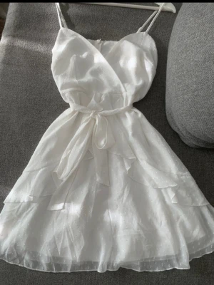 Vit sommarklänning med volanger - Söt vit klänning med tunna axelband, omlottliv och knytband i midjan. 