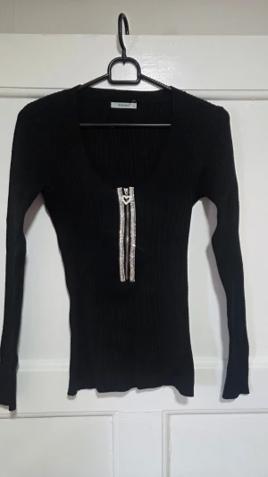 Svart ribbad topp med dragkedja - Snygg svart ribbad topp från Enzoria med lång ärm och en glittrig dragkedja framtill som har ett hjärtformat handtag. Stretchigt material som sitter tight och markerar figuren. Perfekt för dig som gillar detaljer och vill sticka ut lite extra.