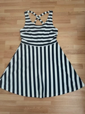 Randig kort klänning med korsade band - Snygg kort klänning som ny med svartvita ränder och korsade axelband i ryggen. Klänningen har ett figurnära liv med horisontella ränder och en utställd kjol med vertikala ränder. Perfekt för sommarens alla tillfällen! Storleklapp bortklippt, men är storlek M, bröst 90cm. Använd en gång.