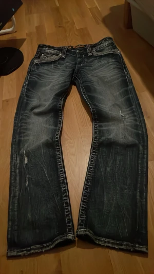 Mörkblåa rock revival jeans - Säljer ett par mörkblåa jeans med slitningar och snyggt broderade bakfickor. Jeansen har kontrastsömmar och en tvättad look med tydliga whiskers och slitna detaljer.