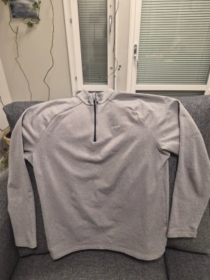 Grå turtle neck half zip tröja från Odlo XXL - Mjuk och skön grå tröja från Odlo med half zip-dragkedja framtill. Tröjan har lång ärm och hög krage, perfekt för kyliga dagar. Enkel och stilren design med diskret logga på bröstet. Passar till träning eller som mellanlager.