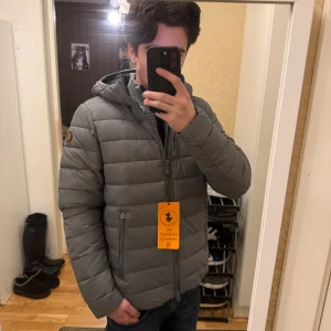 Save The Duck Lightweight jacka - Säljer en grå tunn jacka från Save The Duck med huva och dragkedja framtill. Jackan är tillverkad av 100% återvunnet syntetmaterial och har två sidofickor med dragkedja. Diskret logga på ärmen och coolt orange tag. Perfekt för vår och tidig sommar 🍀