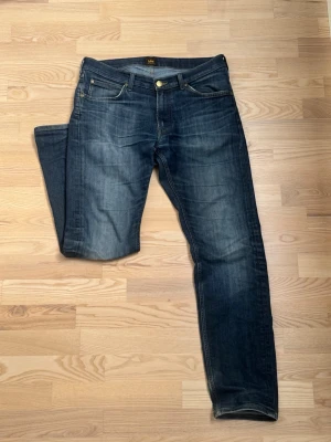 Mörkblå jeans från Lee - Snygga mörkblå jeans från Lee med klassisk femficksdesign och guldfärgad knapp. Jeansen har en lätt tvättad look och Lee-lapp bak i midjan. Perfekta för dig som gillar en tidlös och avslappnad stil. Slitningar och fades från användning, se bilder. Storlek W33L32